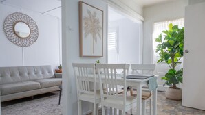 Ein Wohnzimmer mit grauem Sofa, weißem Esstisch mit Stühlen, einem gerahmten Bild an der Wand und einer großen, grünen Pflanze.
