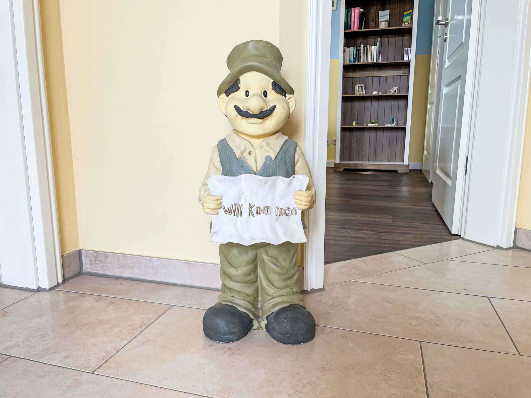 A statue of a man holding a sign that says "Willkommen" in a hallway with a tiled floor and a bookshelf in the background.