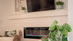 A modern living room with a flat-screen TV mounted above a fireplace, wicker furniture, and potted plants. - Welcome to The Florida Rooms- Your Relaxing Retreat Near the Gulf! New Pool Spa! (Bradenton)