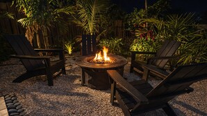 A circular fire pit with flames, surrounded by two wooden chairs and a bench, set on gravel with tropical plants in the background. - Welcome to The Florida Rooms- Your Relaxing Retreat Near the Gulf! New Pool Spa! (Bradenton)