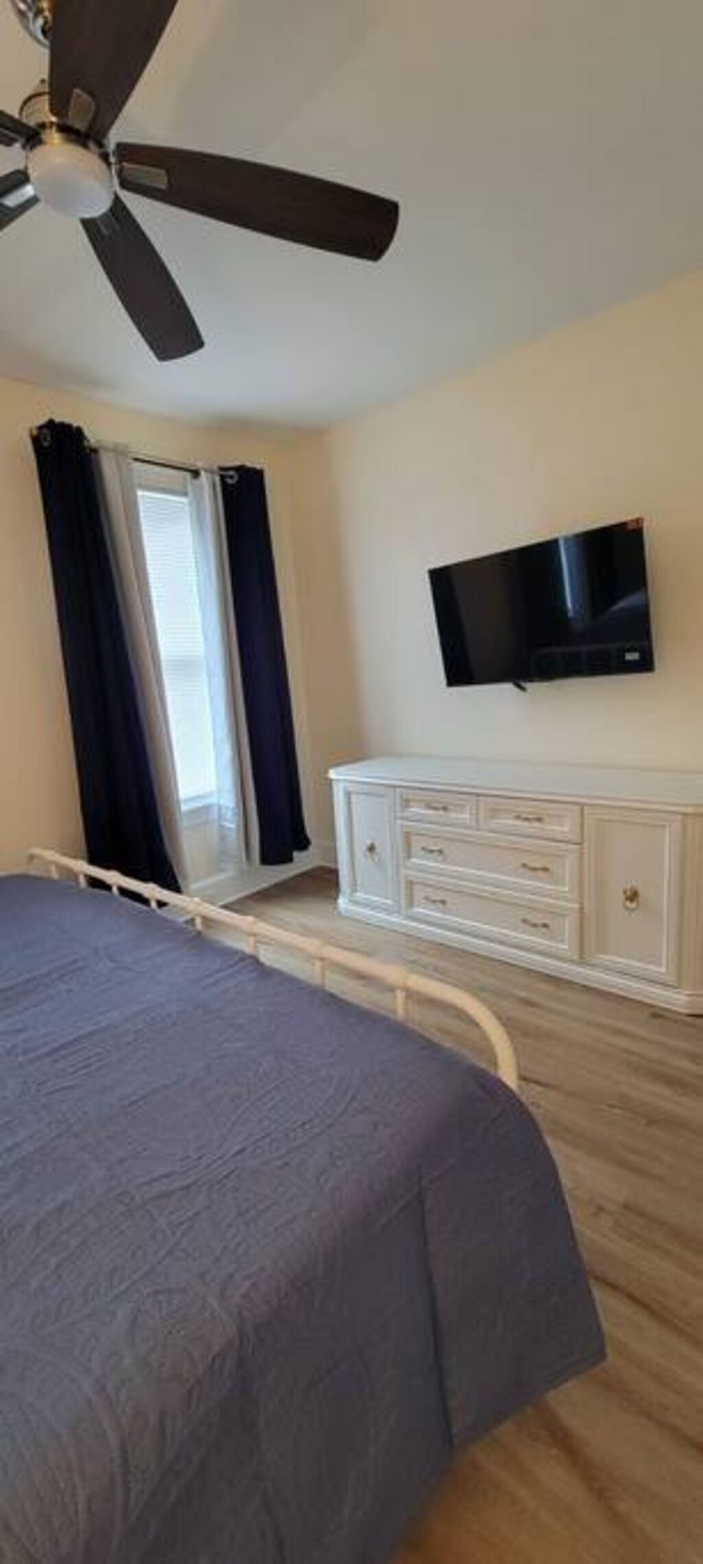 Ein Schlafzimmer mit Bett, Deckenventilator, an der Wand montiertem Fernseher, Kommode und Fenster mit Vorhängen.