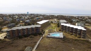 An aerial view of a coastal residential area with multiple apartment buildings, a swimming pool, and a lighthouse in the distance.