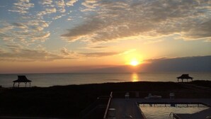 A sunset view from a deck overlooking a beach with lounge chairs and a small structure.