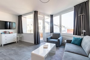 Un salon moderne avec un canapé, un fauteuil et une table basse. Un téléviseur est fixé au mur, il y a une petite table d’appoint et une fenêtre avec des rideaux.