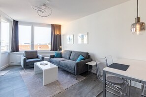 A modern living room with a grey sofa, two armchairs, a white coffee table, and a glass dining table with chairs. There is a large window with curtains, a radiator, and a floor lamp.