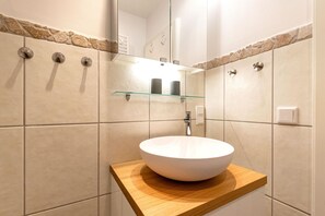 A modern bathroom with a round white sink on a wooden countertop, beige tiles, and a glass shelf above the sink. - Sandbank Domizil in Haus Seeblick - view of the North Sea! - Lake view (Borkum)