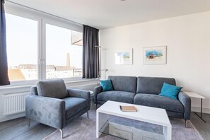 Un salon moderne avec un canapé gris, deux fauteuils, une table basse blanche et une vue sur les immeubles à travers de grandes fenêtres.