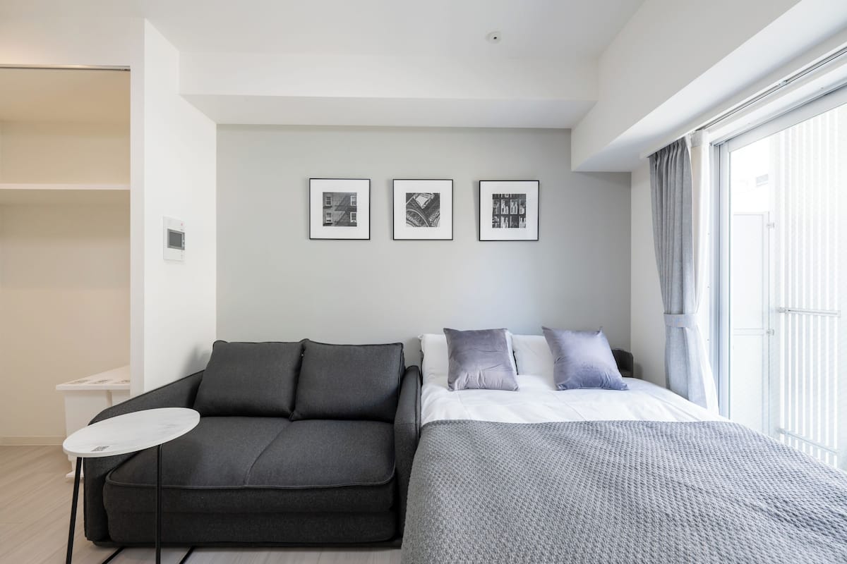 A modern hotel room with a grey sofa, a small round table, a bed with grey bedding, and three framed pictures on the wall.