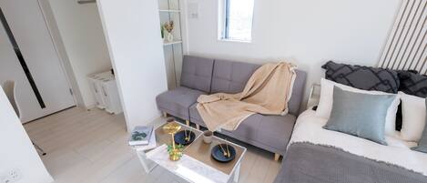 A modern, well-lit room with a grey sofa, a white bed with grey bedding, a small white coffee table with decorative items, and a window with blinds.