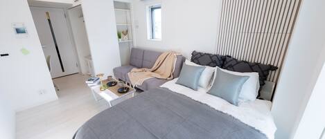 Un dormitorio moderno con una cama grande, un sofá gris, una mesita con una lámpara y una ventana con persianas.