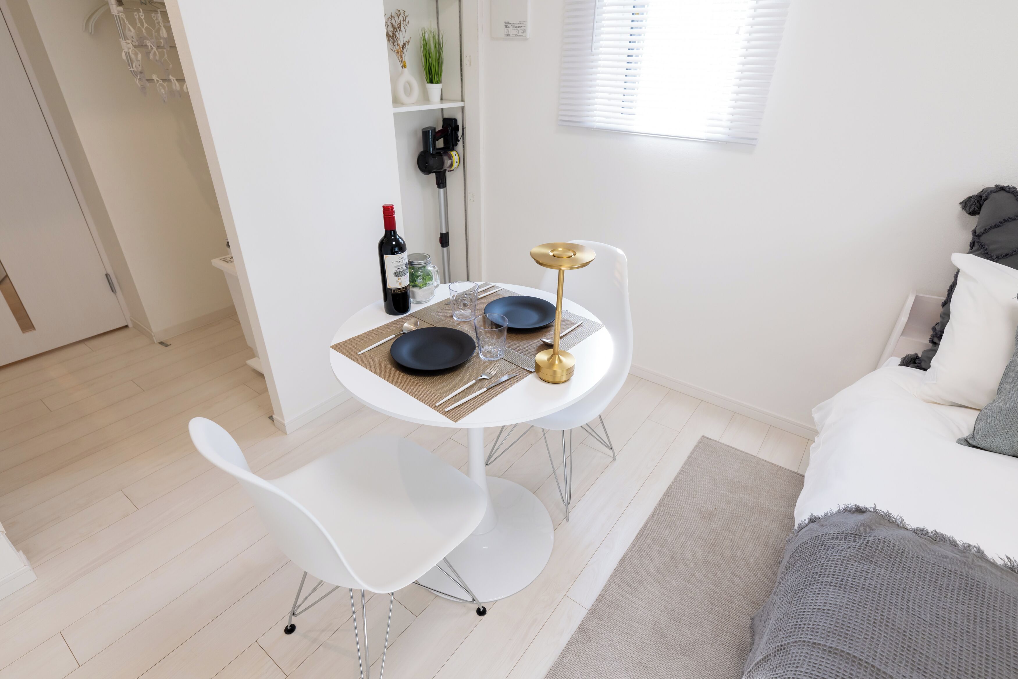A modern, well-lit room with a dining table set for two, a white chair, and a bed with a gray blanket.