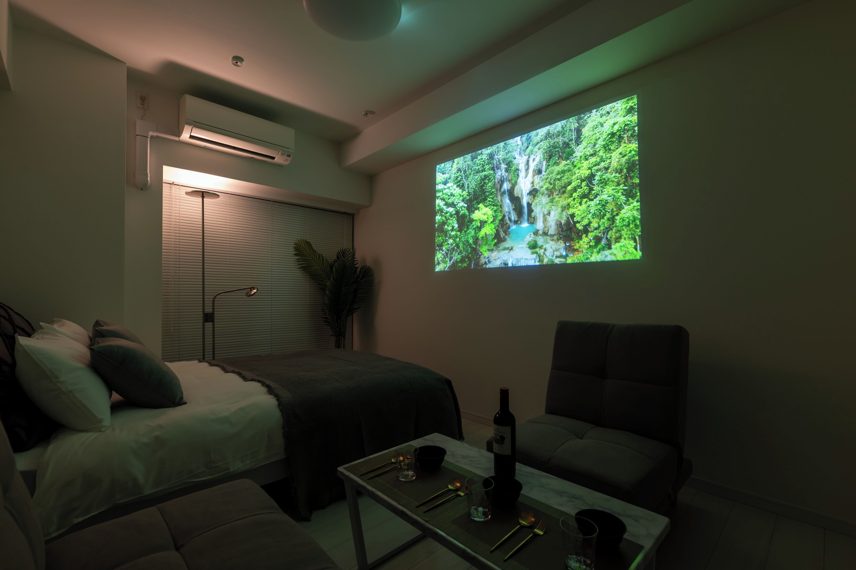 A modern bedroom with a bed, a sofa, a coffee table with a bottle and glasses, and a large screen displaying a waterfall.