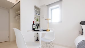 A modern, minimalistic room with a white dining table and chairs, a small kitchen area, and a window with blinds.