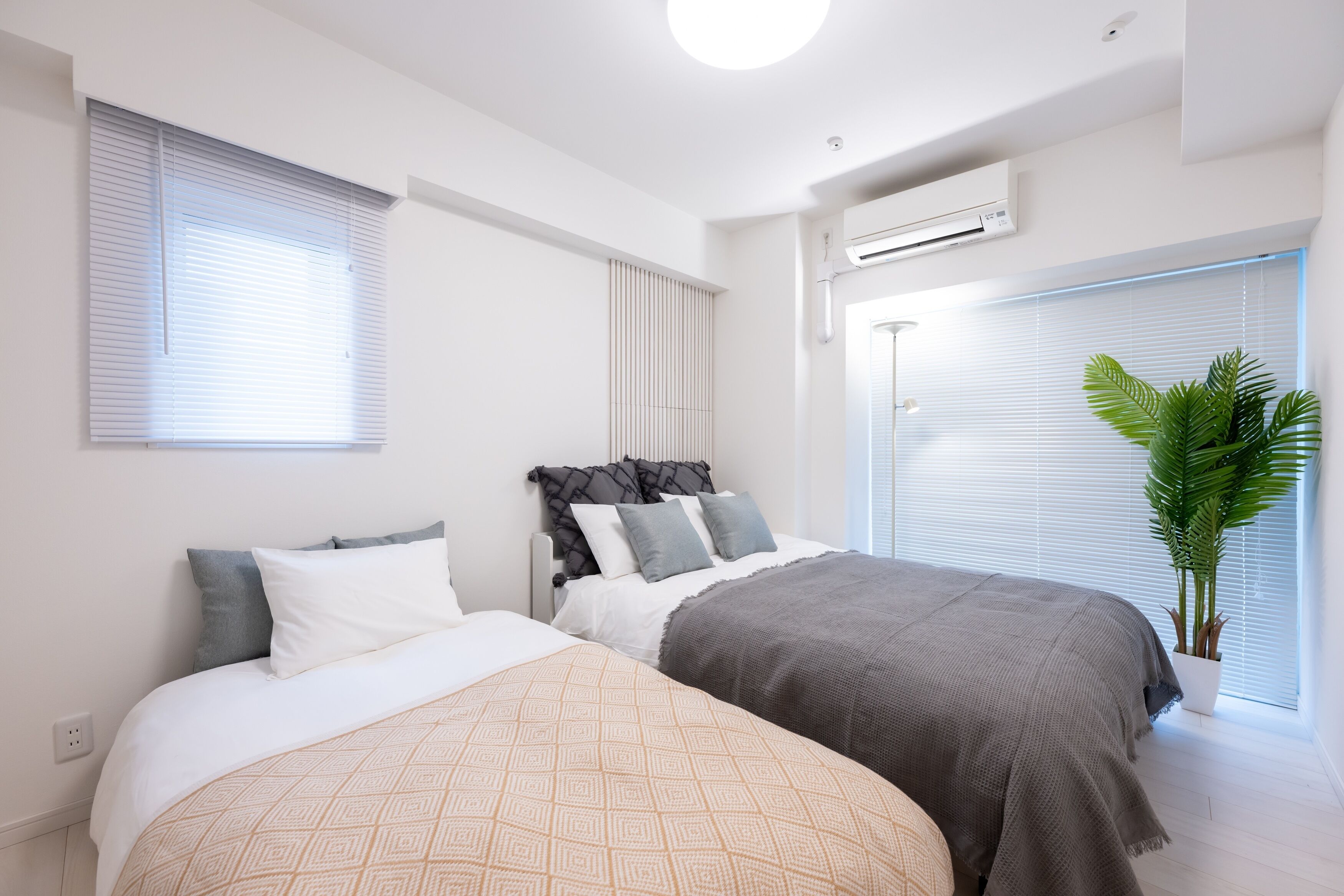 A modern bedroom with two beds, a large window with blinds, an air conditioning unit, and a potted plant.