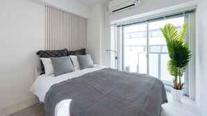 A modern bedroom with a large bed, a potted plant, and a window with a view of buildings.