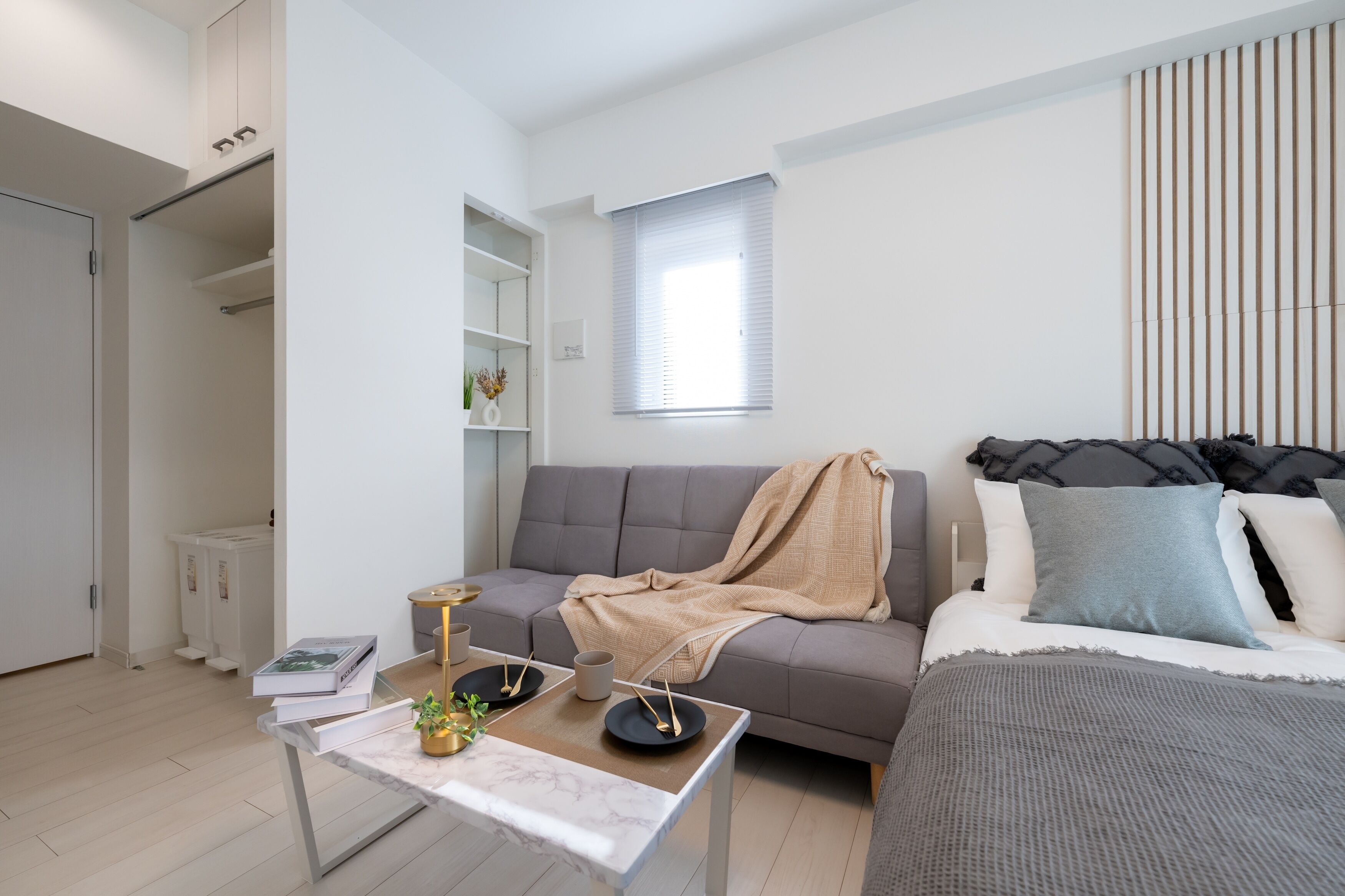 A modern, minimalist bedroom with a grey sofa, a bed with multiple pillows, a marble coffee table, and a small side table with a plant.
