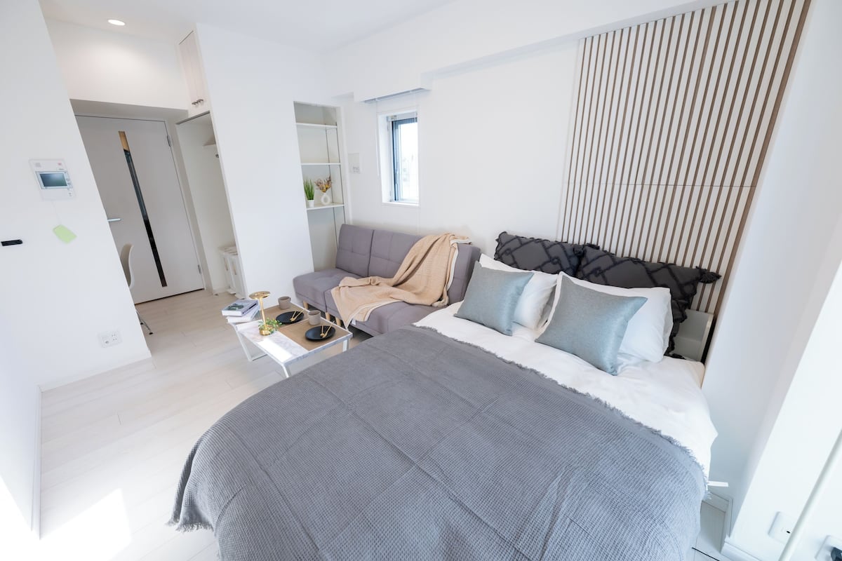 A modern bedroom with a large bed, a sofa, and a small table. There is a window with blinds and a door leading to another room.