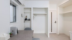 A modern, minimalist room with a grey sofa, a small table with a laptop, a window with blinds, and a white wardrobe with a door.