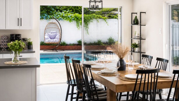 A modern kitchen with white cabinets, a dining area with a wooden table and black chairs, a hanging chair by the pool, and a glass door leading to a garden with plants and a view of the pool. - Spacious 4BR Home w/ Cozy Fireplace, Pool & Easy Access to LA Attractions (Calabasas)