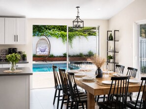 Eine moderne Küche mit weißen Schränken, ein Essbereich mit Holztisch und schwarzen Stühlen, ein Hängesessel am Pool und eine Glastür, die zu einem Garten mit Pflanzen und Blick auf den Pool führt.