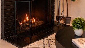 A fireplace with a visible fire, a potted plant, and a round table with a book and a beaded necklace. - Spacious 4BR Home w/ Cozy Fireplace, Pool & Easy Access to LA Attractions (Calabasas)