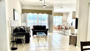 Un salon moderne avec un canapé, un fauteuil et une table basse, qui donne sur un coin repas avec des chaises et une cuisine en arrière-plan.