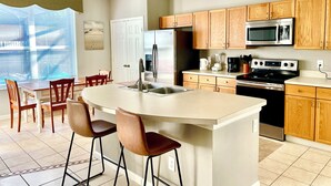 Une cuisine moderne avec des meubles en bois, des appareils électroménagers en acier inoxydable, un îlot central doté de tabourets et un coin repas avec des chaises.