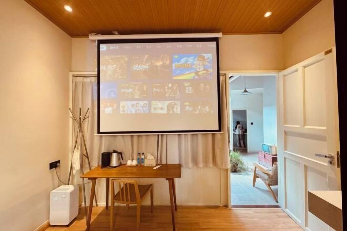 Une pièce dotée d’un plafond en bois, d’un écran de projection affichant diverses images, d’une table en bois avec une chaise, et d’un purificateur d’air blanc.