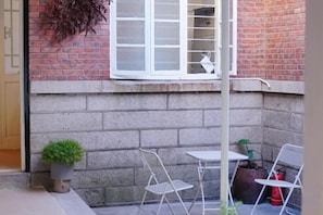 Un patio lateral con una mesa y sillas blancas, una planta en maceta y una ventana con vista a un muro de ladrillo.