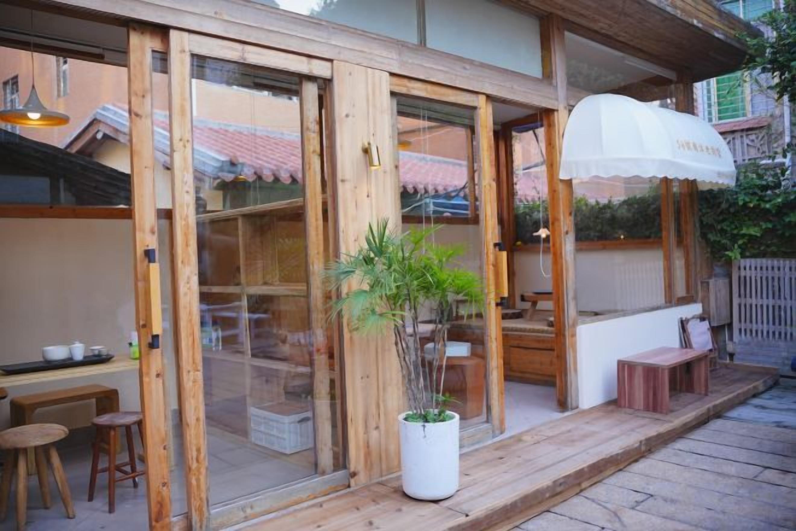 A wooden exterior with large glass doors, a potted plant, outdoor seating, and a white umbrella.