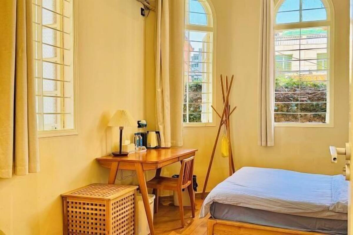 Une pièce comprenant un lit, une table en bois, une lampe et une fenêtre donnant sur des immeubles.