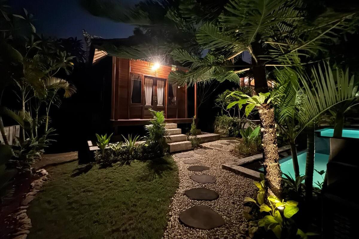 Vista nocturna de una casa con fachada de madera, un camino de piedra y una piscina.