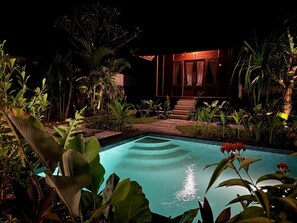 Une vue nocturne d’une piscine entourée d’une végétation tropicale luxuriante, avec un bâtiment en bois en arrière-plan.
