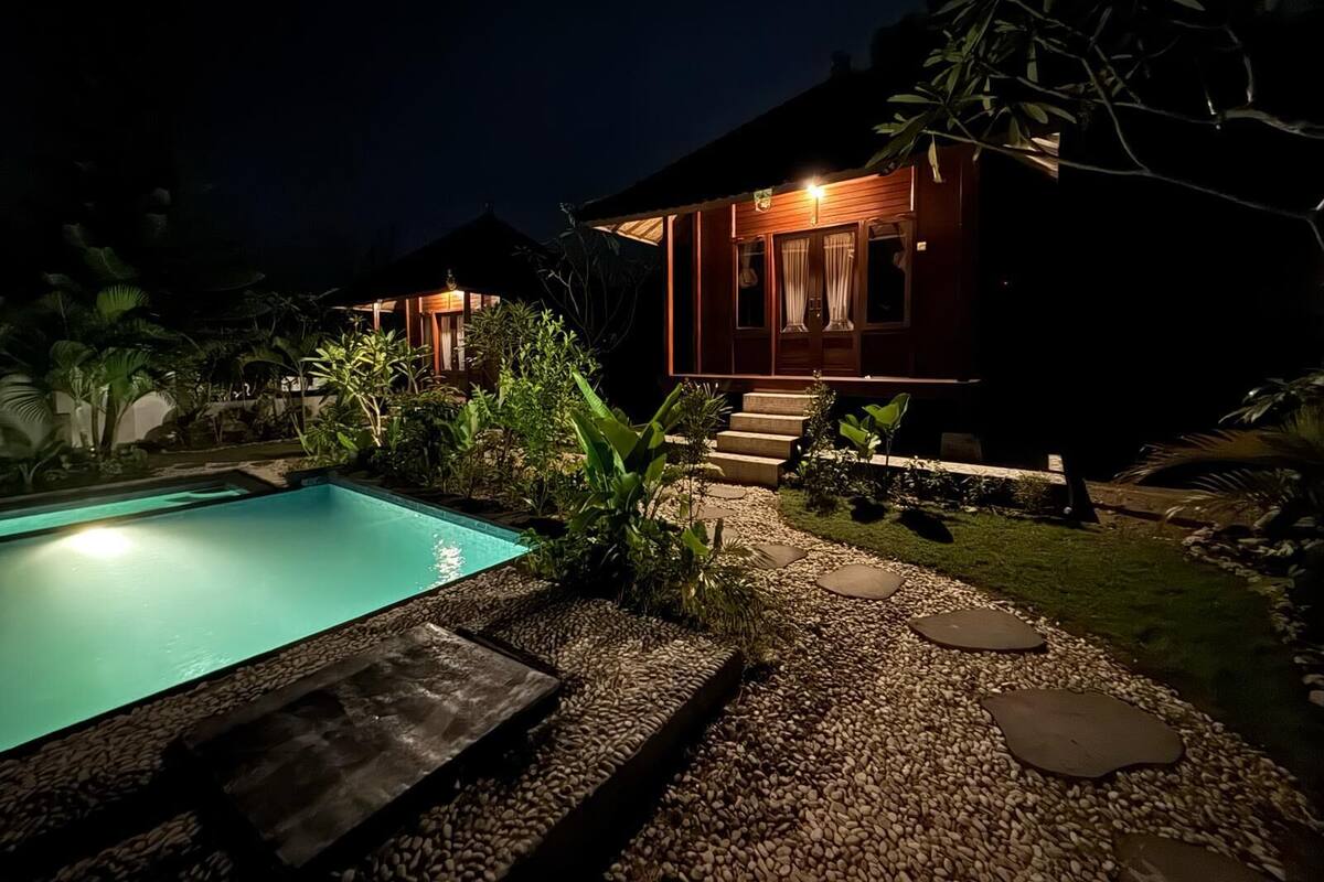 Vista nocturna de una casa de dos pisos con piscina, rodeada de plantas tropicales y un camino de piedra.