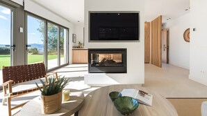 A modern interior with a large sliding glass door, a flat-screen TV mounted on the wall, a wooden bench, and a dining table with a bowl.