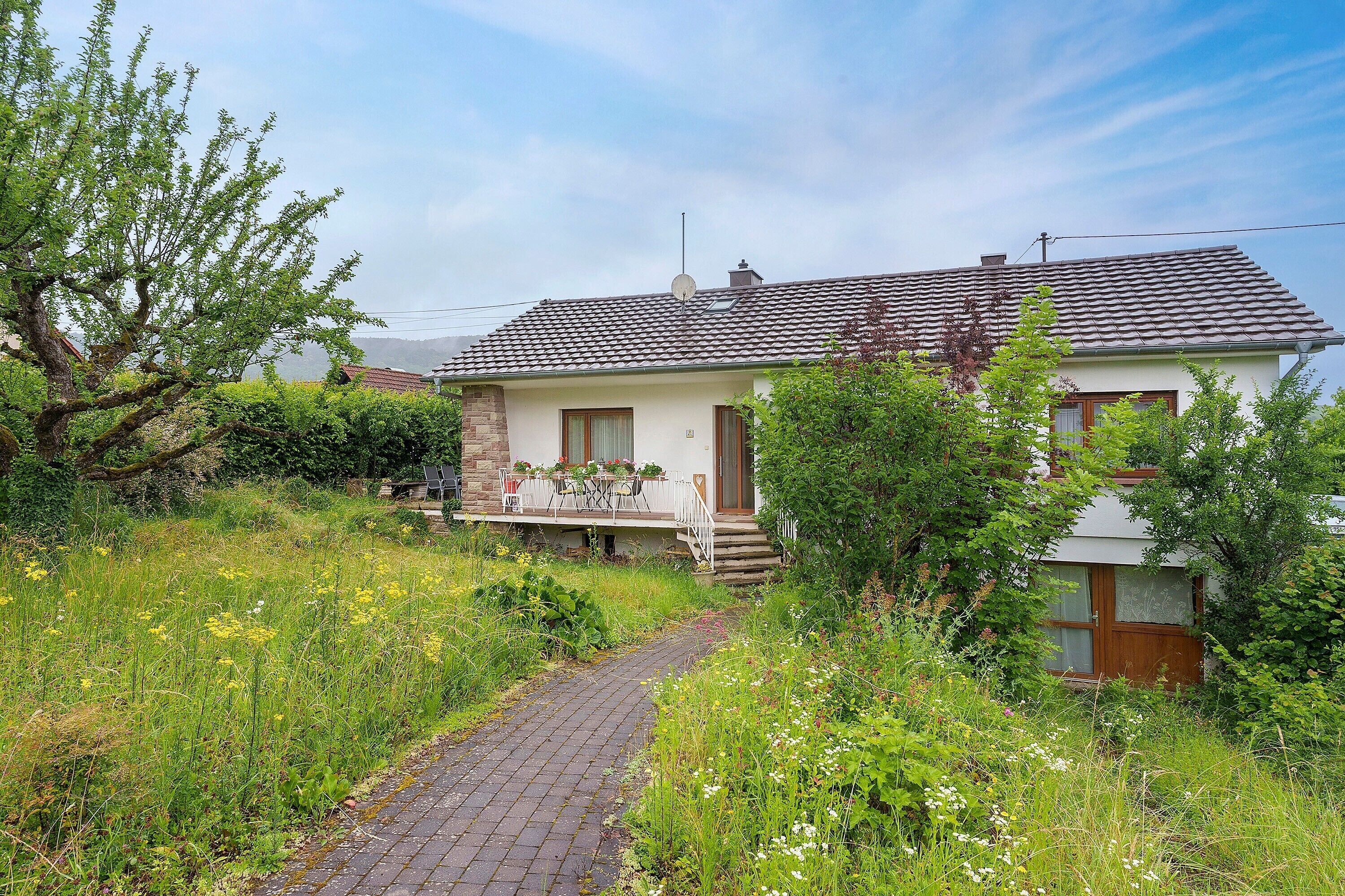 A house with a paved pathway leading to the entrance, surrounded by lush greenery and a clear sky.