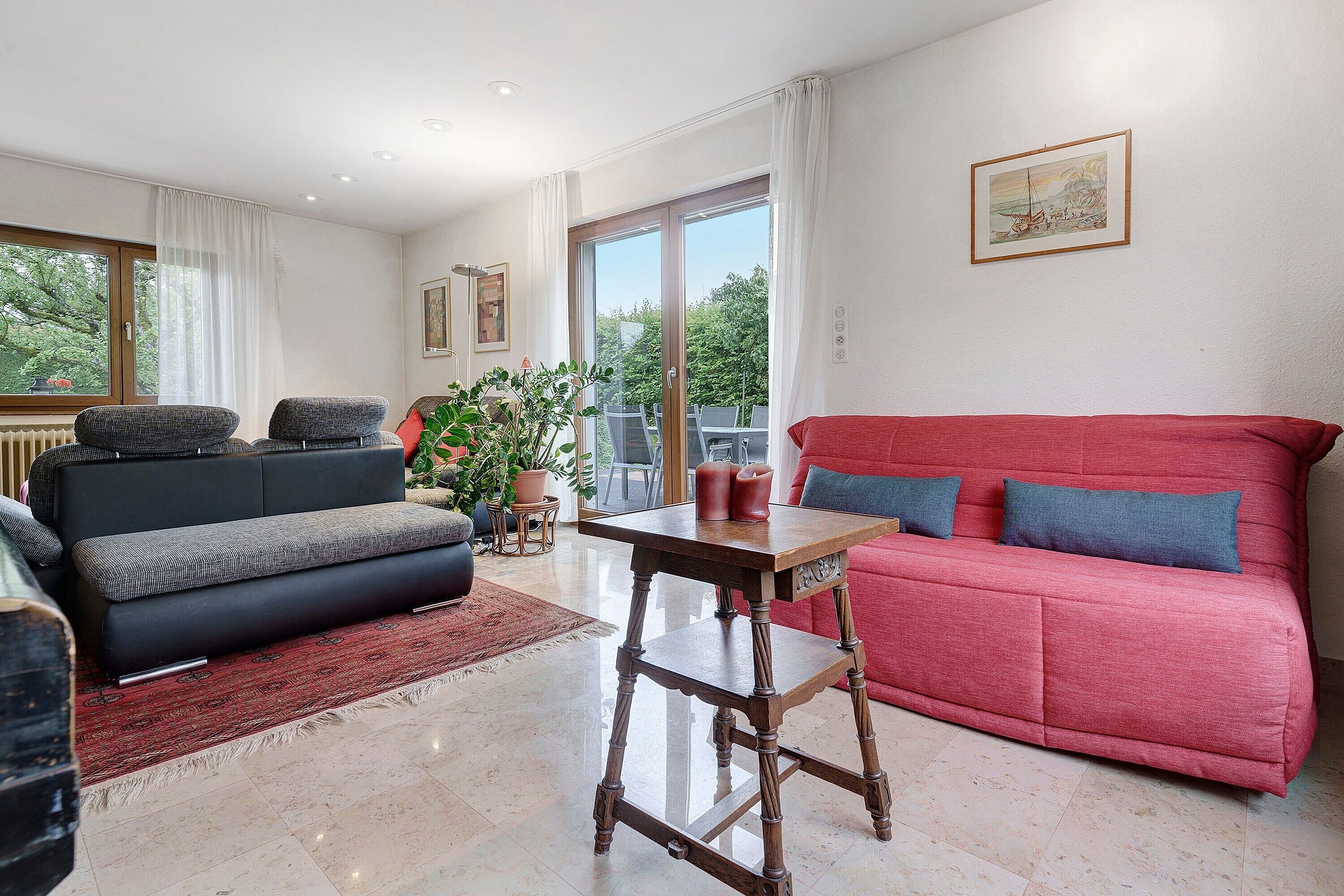 A living room with a sofa, a table, and a chair. There is a window with curtains, a plant, and a painting on the wall.