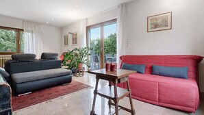 A living room with a sofa, a table, and a chair. There is a window with curtains, a plant, and a painting on the wall. - Holiday Home 'Le Château D'Eau' with Private Terrace, Private Garden and Wi-Fi (Wissembourg)