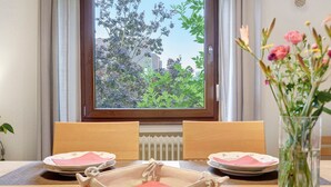 A dining table with a vase of flowers, a window with a view of greenery, and a radiator below the window. - Holiday Home 'Le Château D'Eau' with Private Terrace, Private Garden and Wi-Fi (Wissembourg)