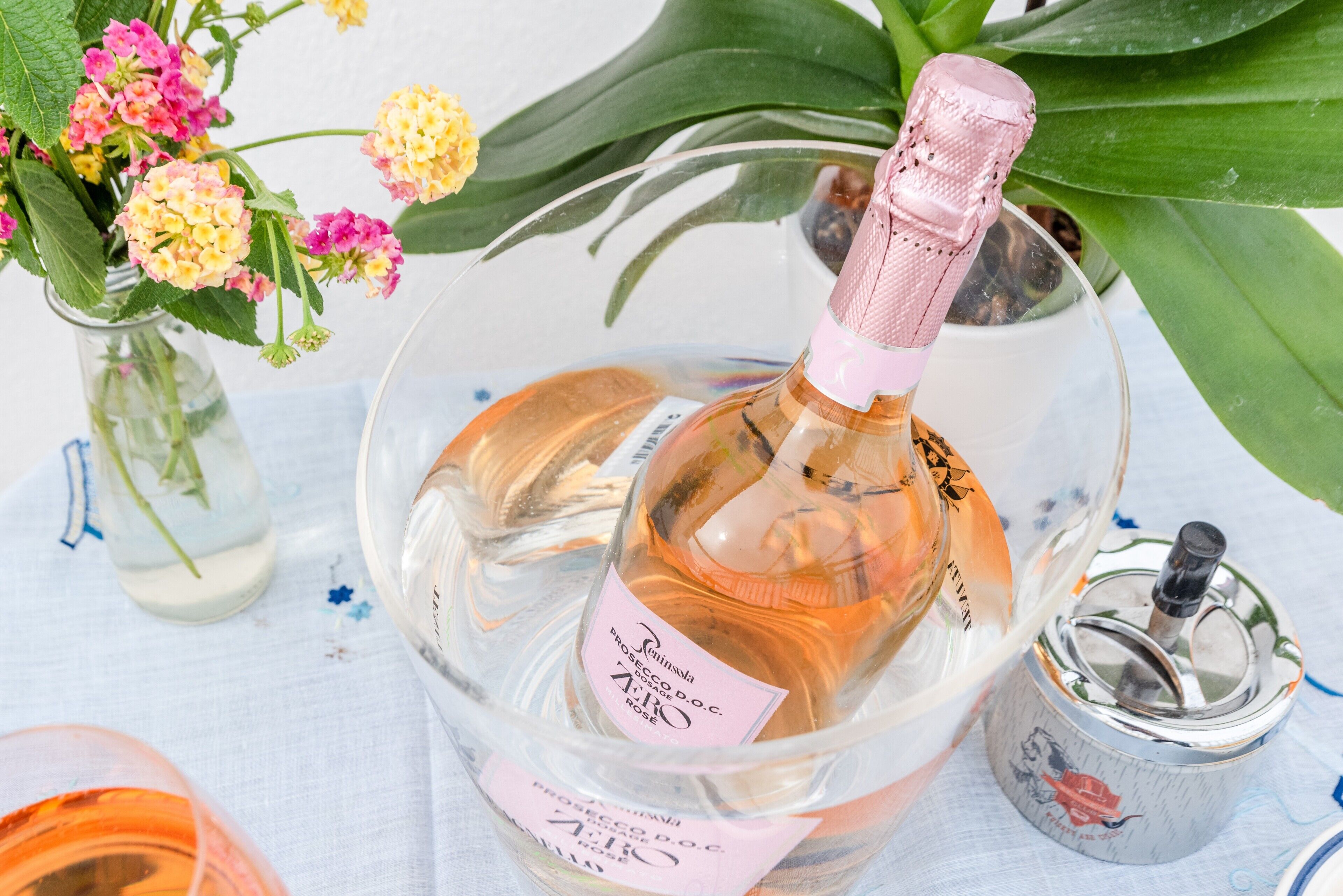 Eine Weinflasche in einem Glasbehälter mit Rosé Korken, ein kleiner Metallbehälter und eine Vase mit Blumen.