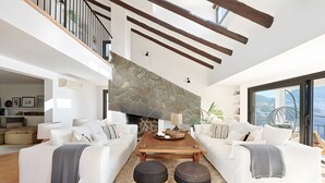 A modern living room with a stone fireplace, white sofas, a wooden coffee table, and a view of the outdoors. - Villa 'Mallorquin Oasis' with Sea View, Wi-Fi and Air Conditioning (Andratx)