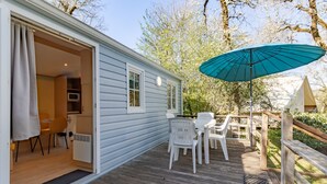 A wooden deck with outdoor seating, a teal umbrella, and a white building with a door open. - Mobile Home "Calvi": Holidays in Dordogne, Shared Pool & Mini-Farm near Sarlat (Bouzic)