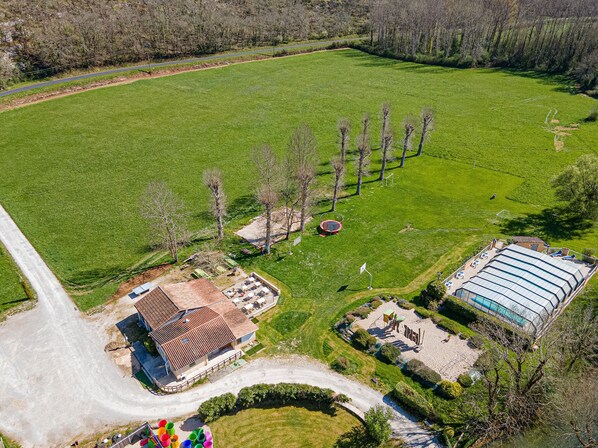 Vue aérienne d’une propriété comprenant un grand terrain verdoyant, une piscine, un bâtiment avec un toit en tuiles et une route asphaltée.