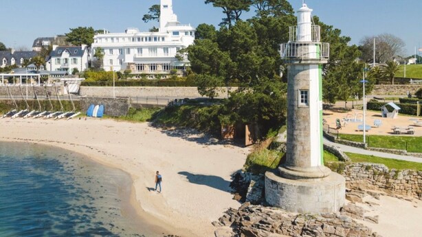 Paisaje costero con un faro, playa y un edificio blanco grande con una torre.