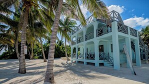 Un edificio azul claro con balcones y una escalera, rodeado de palmeras sobre una playa arenosa.