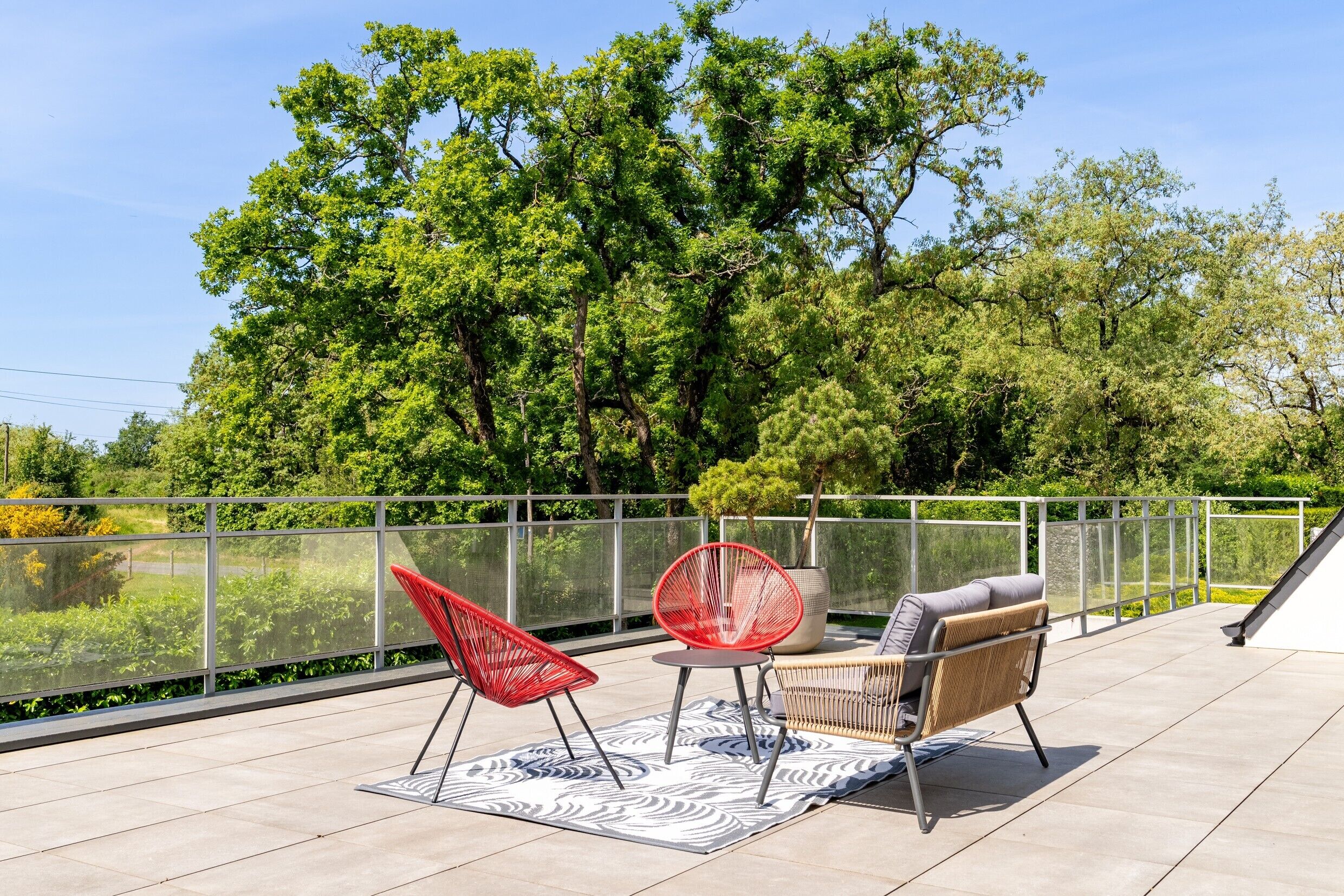 Een terras met buitenmeubilair, een glazen hek en uitzicht op bomen.