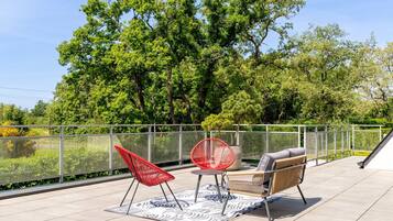 Een terras met buitenmeubilair, een glazen hek en uitzicht op bomen.