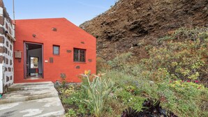 Un bâtiment rouge doté d’une porte et de fenêtres, entouré d’un jardin où poussent différentes espèces de plantes.