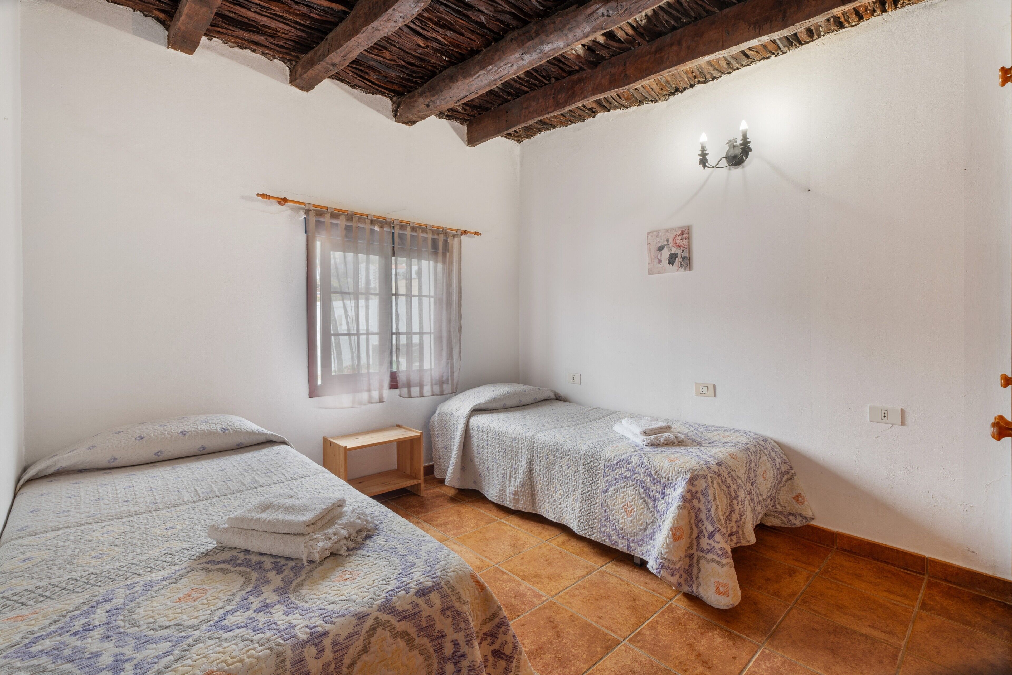 A room with two beds, a small table, a window with sheer curtains, a wall-mounted light fixture, and a rustic ceiling.