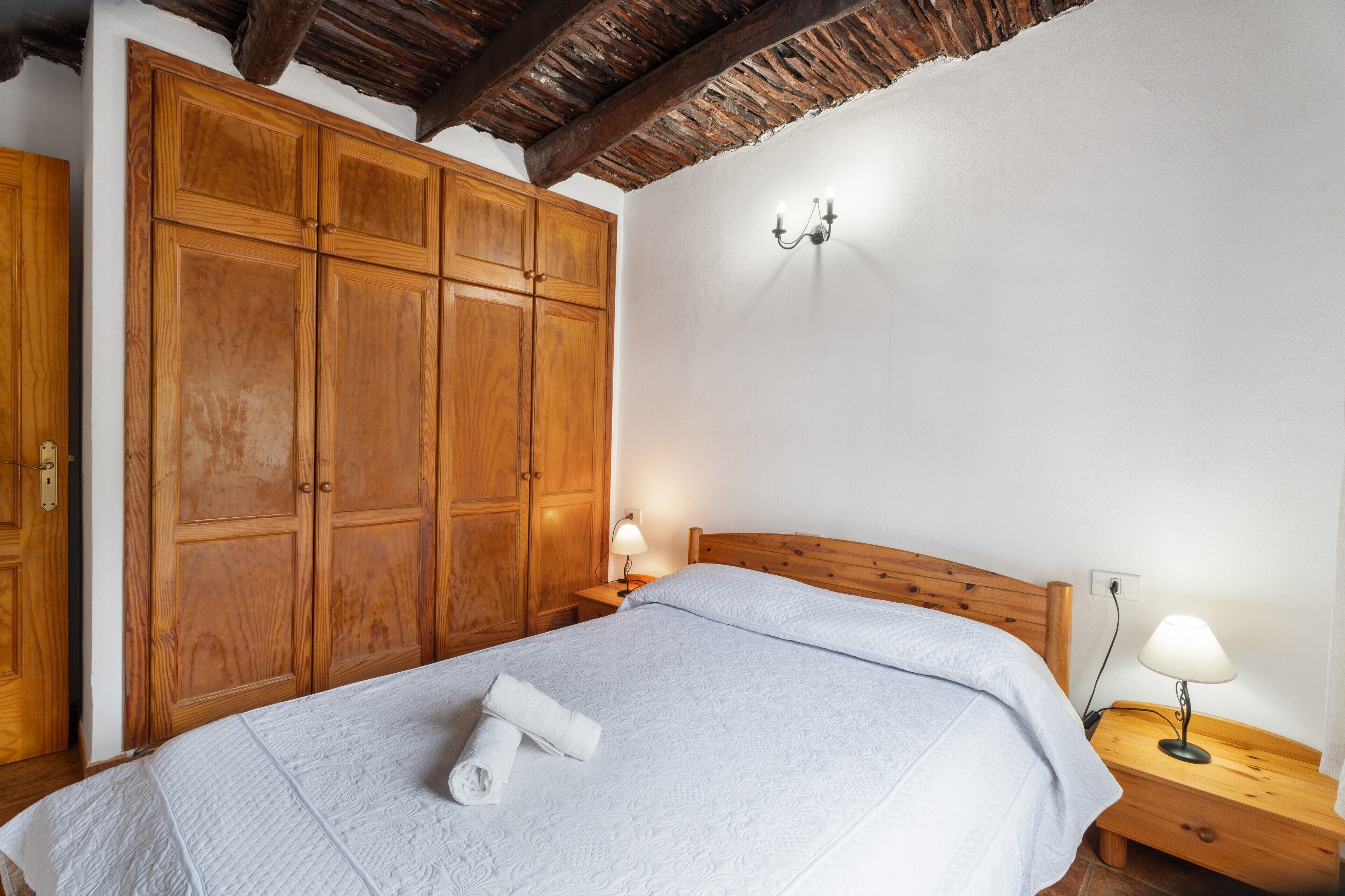 A bedroom with a wooden wardrobe, a bed with a white duvet, a nightstand with a lamp, and a wooden ceiling.
